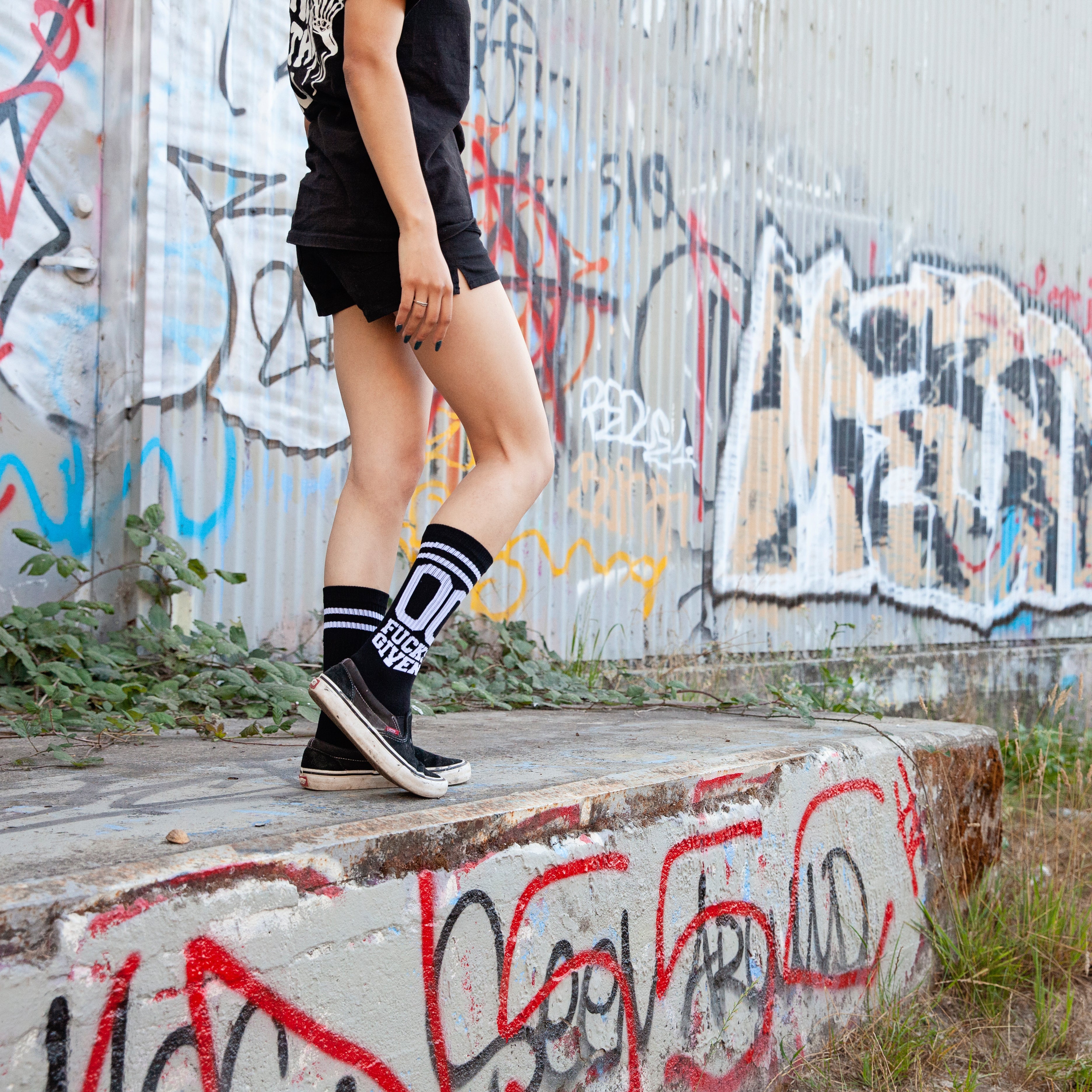Lifestyle shot: Person wearing black '00 Fucks Given' socks and black shoes, sitting on a wall with graffiti background. [lf]