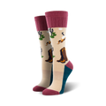 A pair of beige crew socks with maroon cuffs. The socks feature a desert landscape design with cowboy boots, cacti, and birds.