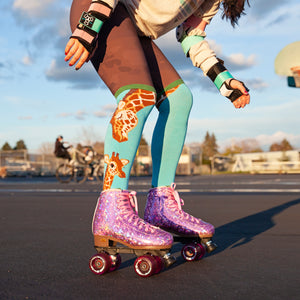 A person wearing sparkly purple roller-skates with pink wheels, blue socks with a giraffe pattern, and protective wrist guards is roller-skating on a paved surface while another person rides a bicycle in the background.