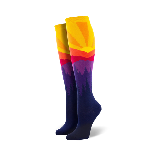 Pair of women's knee-high socks showcasing a vibrant sunset over silhouetted mountains. Colors include yellow, orange, red, purple, and navy blue.