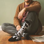 Man wearing black and white crossword crew socks, sitting on a wood floor with a newspaper. [lf]
