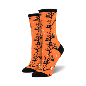 A pair of women's crew socks in bright orange. The socks are covered in a pattern of black owls sitting in bare trees against a background of small white stars and crescent moons.