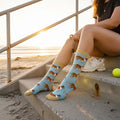 Woman wearing light blue corgi dog crew socks with tan trim, sitting on beach steps at sunset with a tennis ball. [lf]