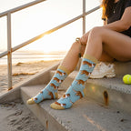 Woman wearing light blue corgi dog crew socks with tan trim, sitting on beach steps at sunset with a tennis ball. [lf]