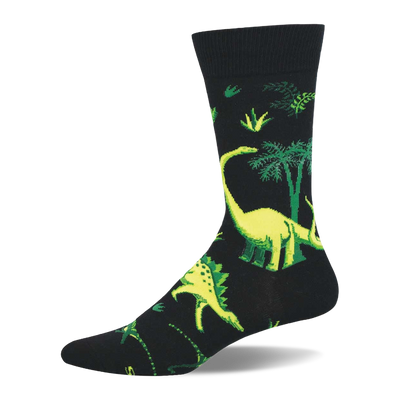 Black crew sock with vibrant yellow-green dinosaurs and green prehistoric trees/foliage. Long-necked and spiky dinosaurs visible.