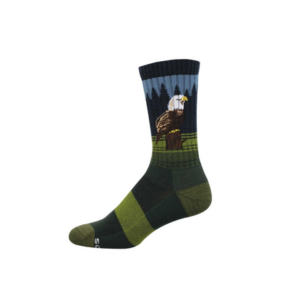 Crew sock with a bald eagle perched on a stump, dark pine trees, and blue sky. Green and dark green stripes on foot.