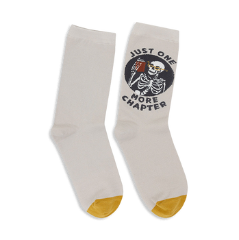 Pair of off-white crew socks with a mustard yellow heel and toe. A circular design shows a skeleton reading a book with the text 'Just One More Chapter'.