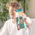 Man laughing, holding up a custom teal crew sock featuring repeated funny faces, indoors with plants. [lf]