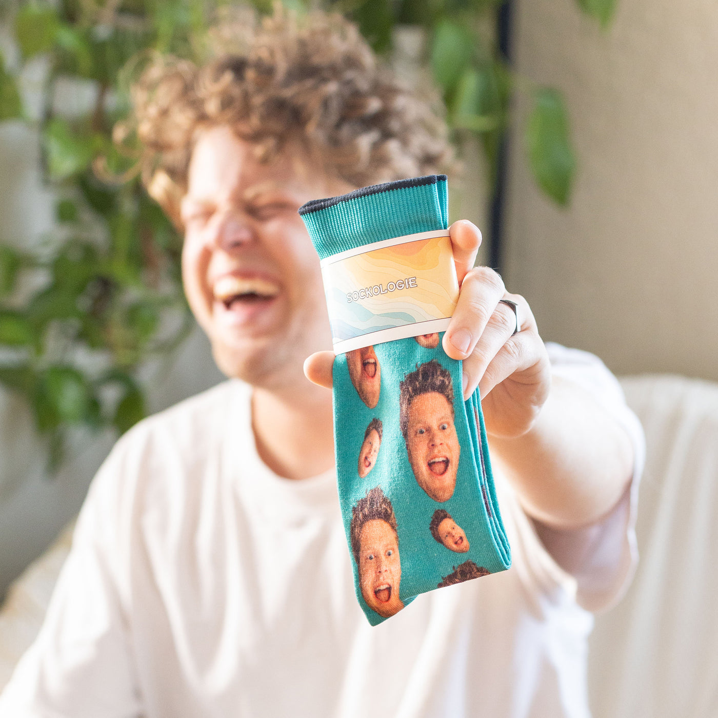 Man laughing, holding up a custom teal crew sock featuring repeated funny faces, indoors with plants. [lf]