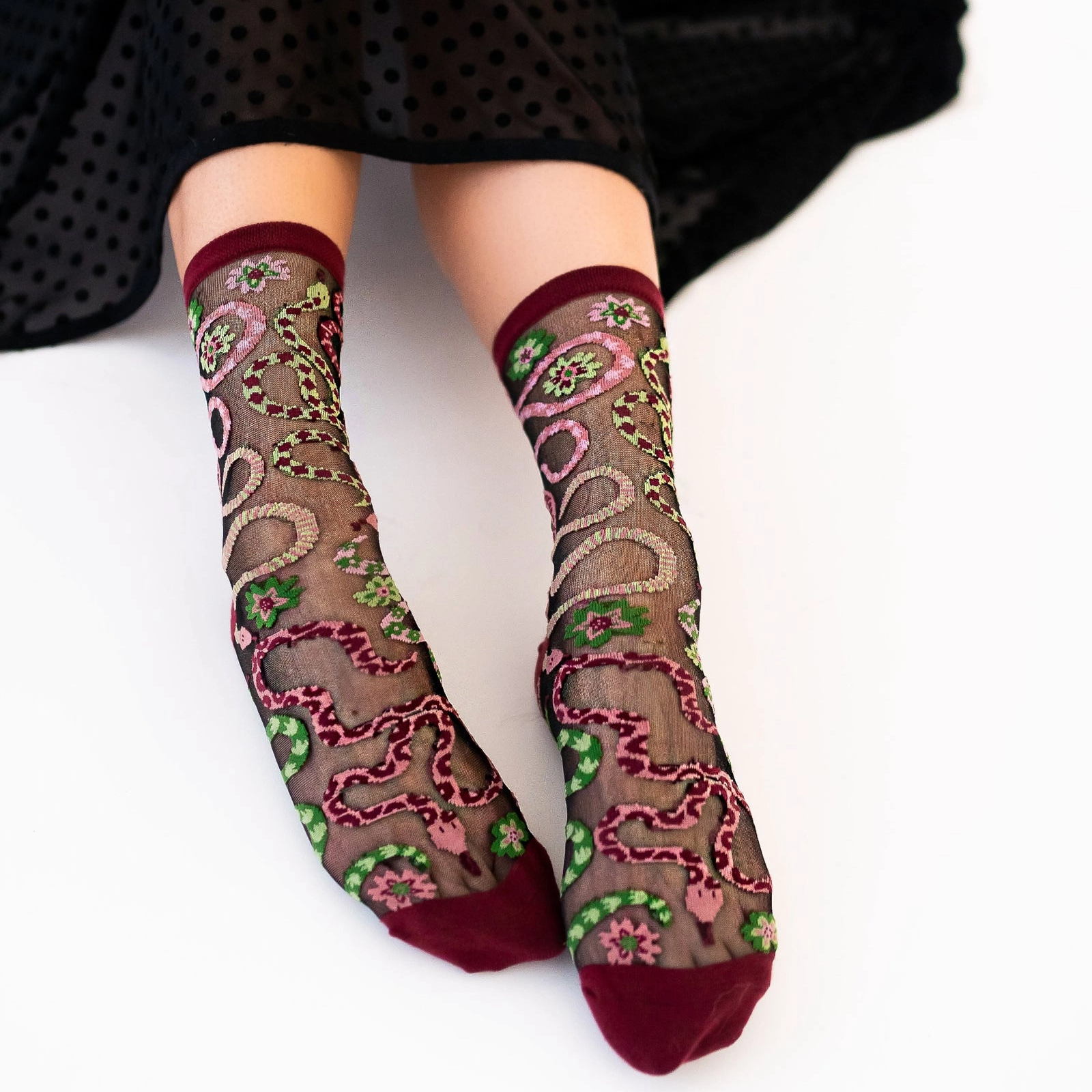 Person sitting wearing sheer snake pattern socks with burgundy trim, a black polka dot skirt draped over their knees.