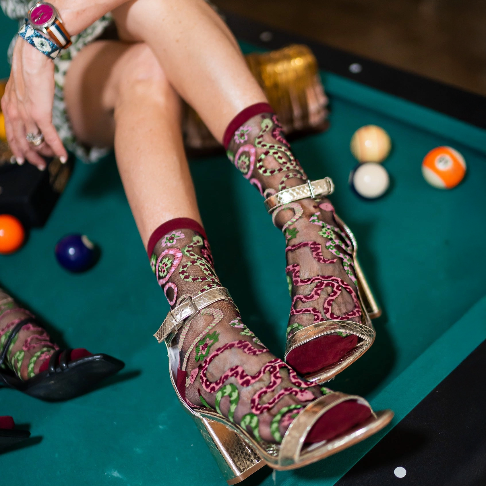 Legs resting on a green pool table wearing sheer snake pattern socks and metallic gold strappy heels next to billiard balls.