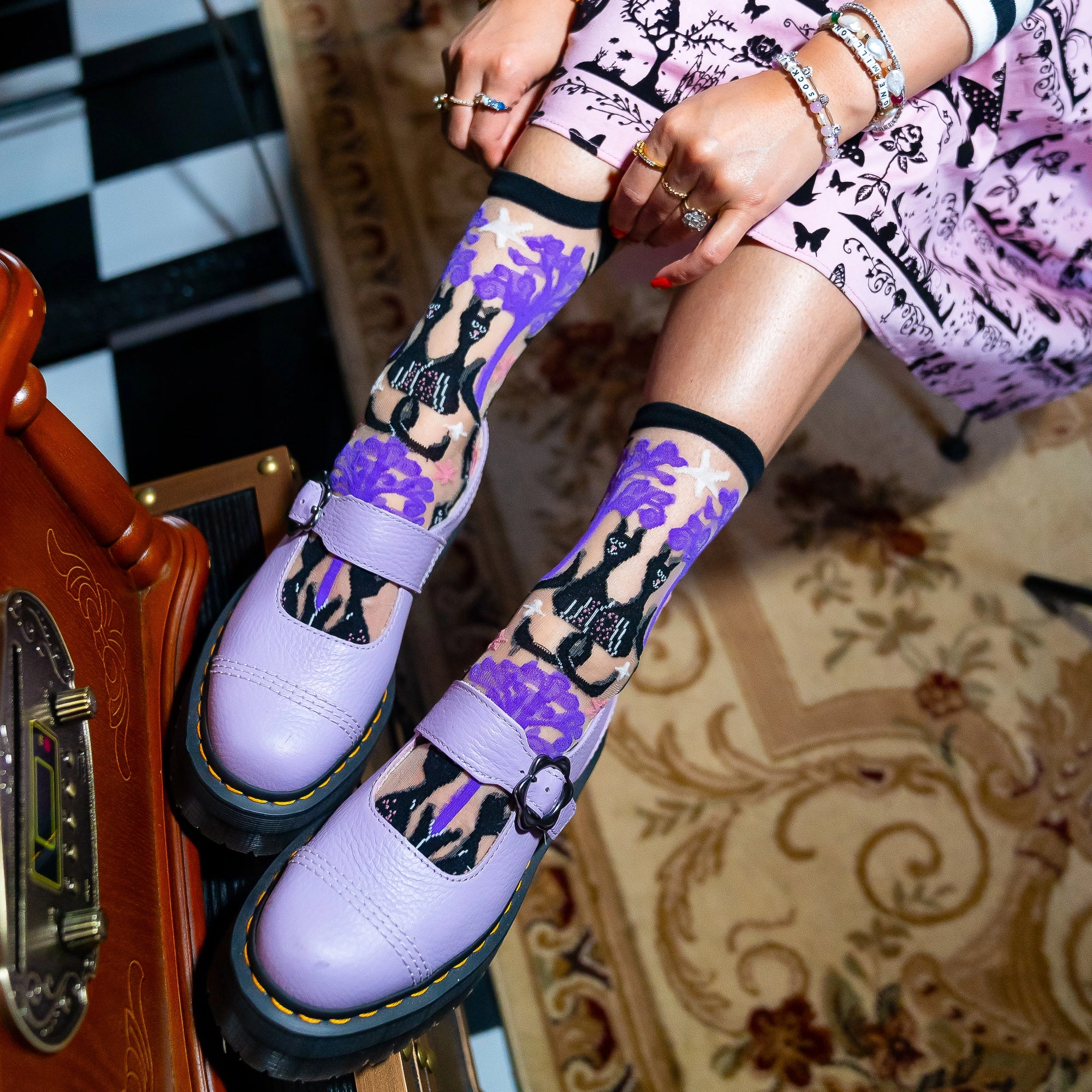 Person wearing sheer cat and purple tree socks with chunky light purple Mary Jane shoes on a patterned rug.