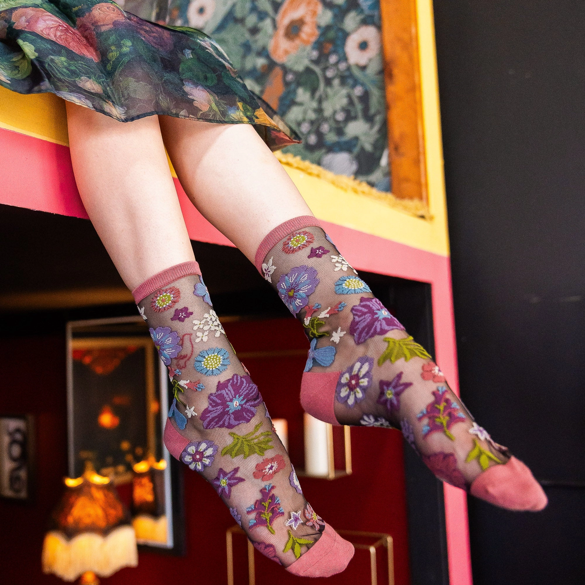 Person legs dangling over a colorful painted ledge showing sheer floral socks and a dark red wall in the background