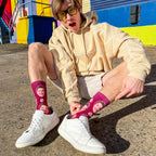 Young man in sunglasses and beige hoodie crouches, highlighting his custom maroon crew sock featuring his face, by a colorful wall. [lf]