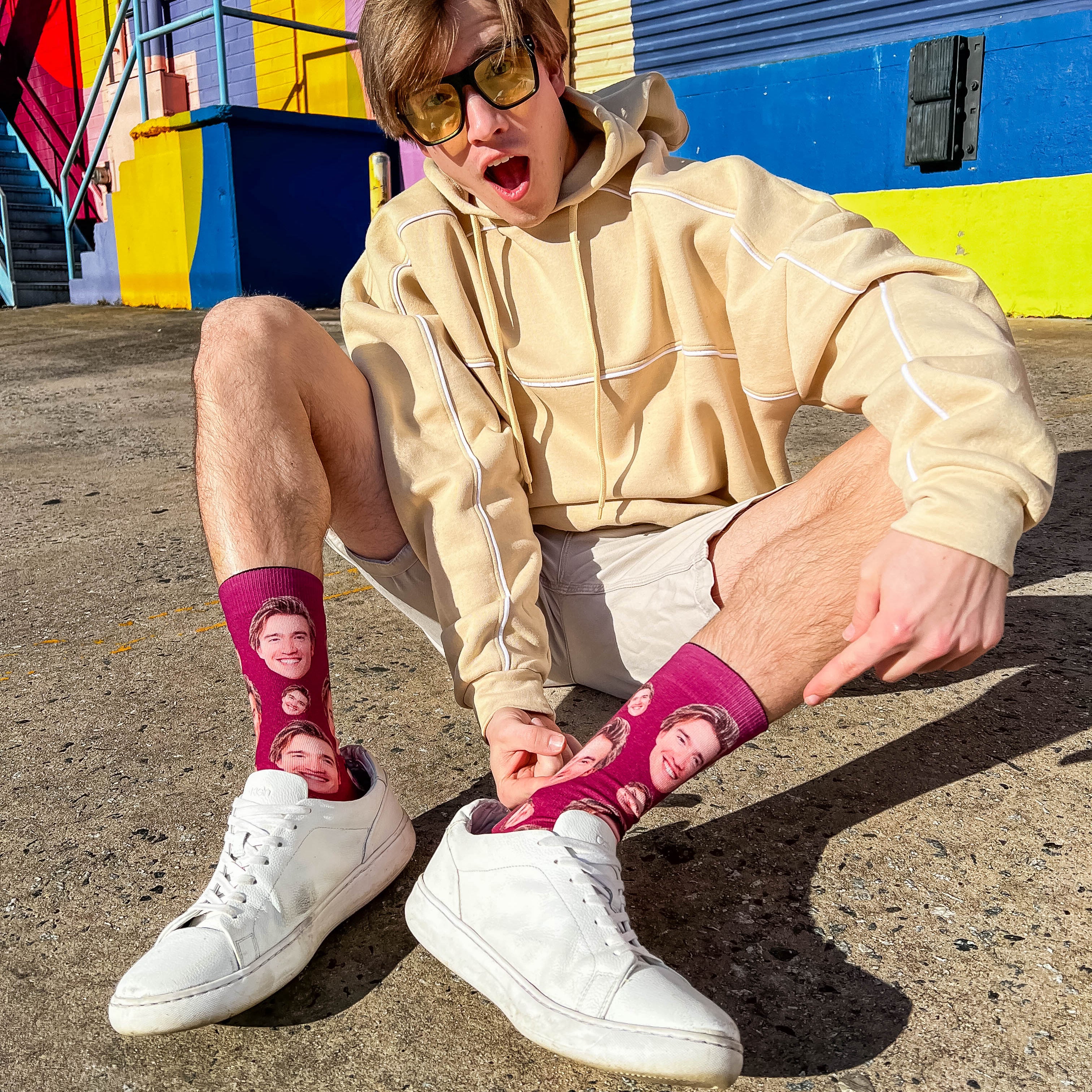 Young man in sunglasses and beige hoodie crouches, highlighting his custom maroon crew sock featuring his face, by a colorful wall. [lf]