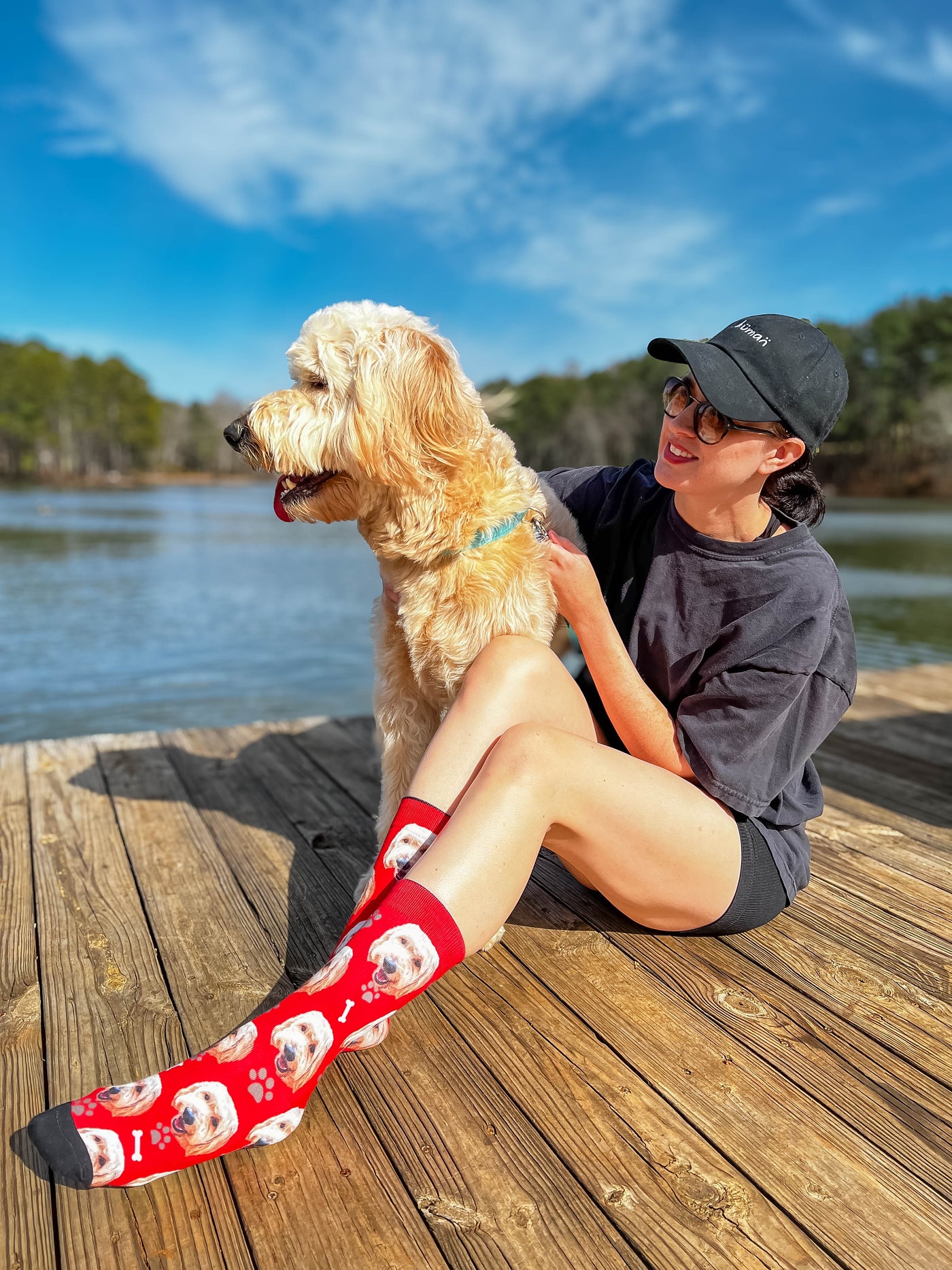 Woman wearing red crew socks with golden doodle faces, bones, and paw prints, petting a dog on a dock. [lf]