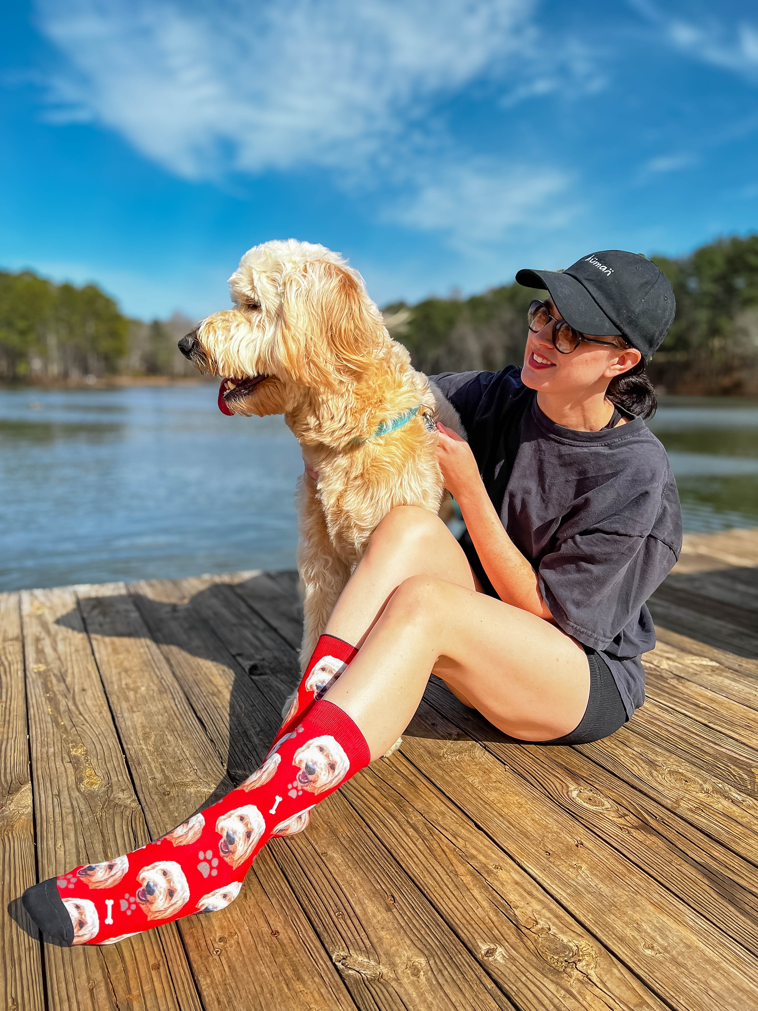 Woman wearing red crew socks with golden doodle faces, bones, and paw prints, petting a dog on a dock. [lf]