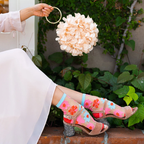 Person holding a round flower purse, wearing sheer floral socks with strappy heels and a white dress on a brick ledge.