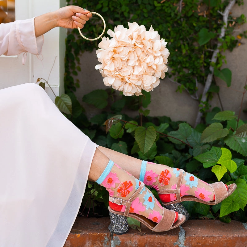 Person holding a round flower purse, wearing sheer floral socks with strappy heels and a white dress on a brick ledge.
