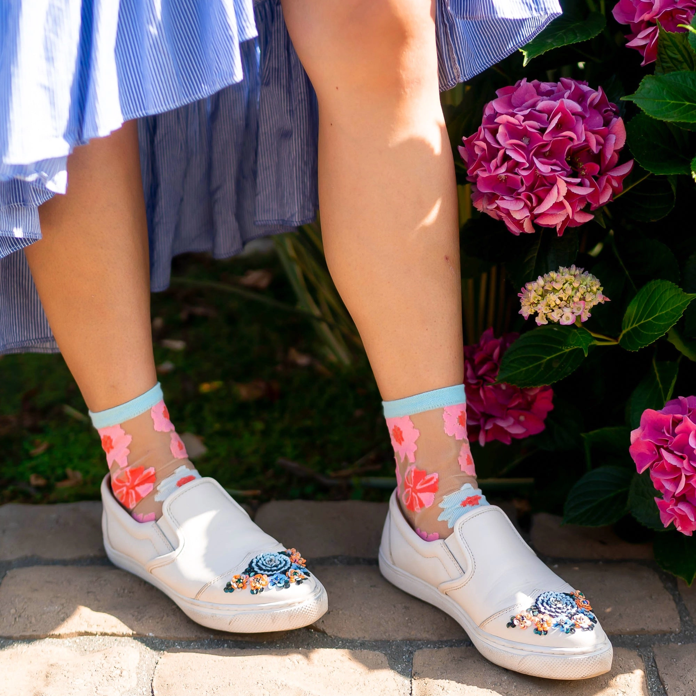 Lower body view of sheer pink and blue floral socks styled with sparkling block heels and a white skirt next to green ivy.