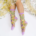 Legs wearing sheer floral socks with purple cuffs, shown against a white floor next to a yellow floral sheer dress.