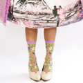 Standing legs wearing sheer vine and flower socks paired with cream vintage heels under a colorful printed skirt.
