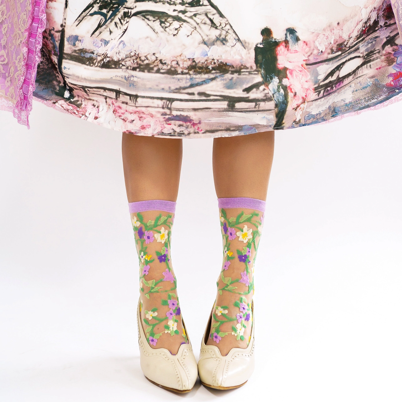 Standing legs wearing sheer vine and flower socks paired with cream vintage heels under a colorful printed skirt.