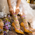 A woman wearing a white lace dress and yellow lace stiletto heels adjusts the purple cuff of her sheer floral sock.