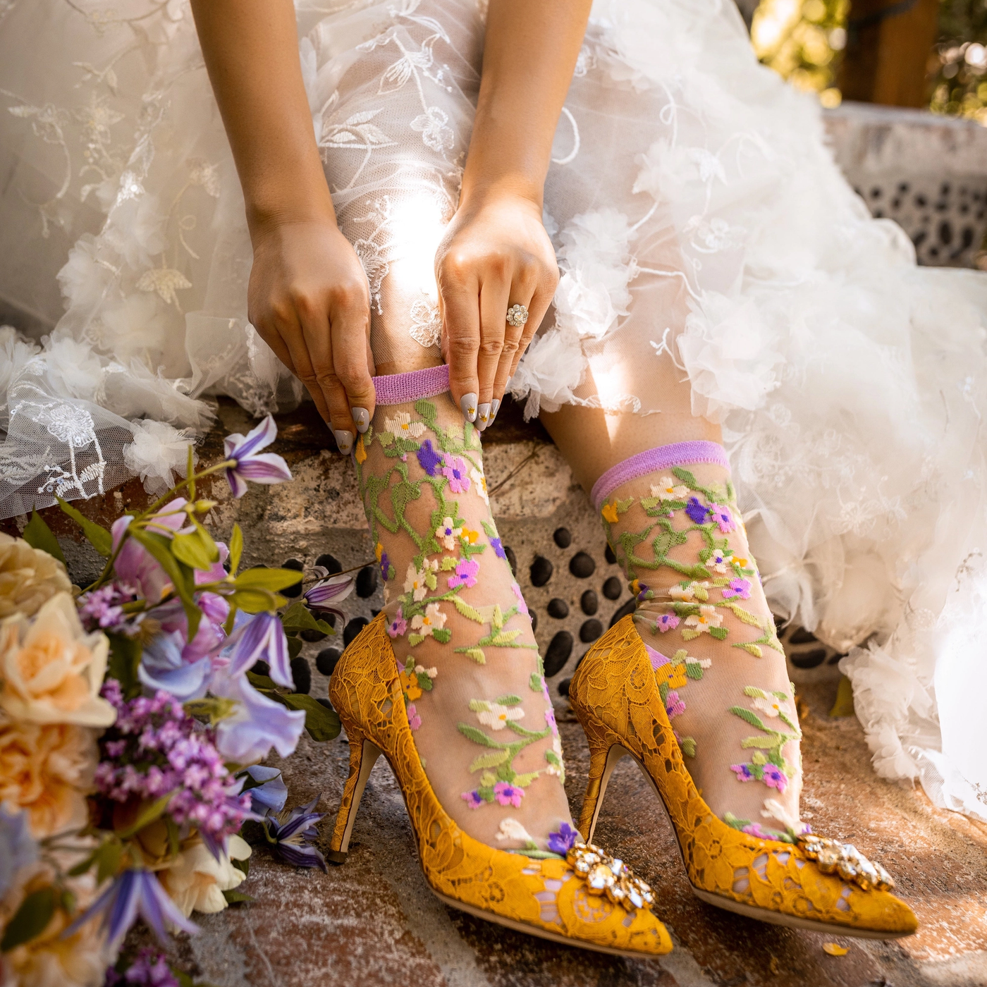 A woman wearing a white lace dress and yellow lace stiletto heels adjusts the purple cuff of her sheer floral sock.