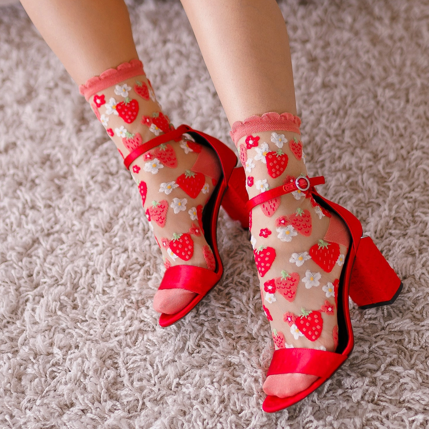 Close-up of feet on a fuzzy white rug, wearing sheer strawberry socks paired with bright red strappy block heels.