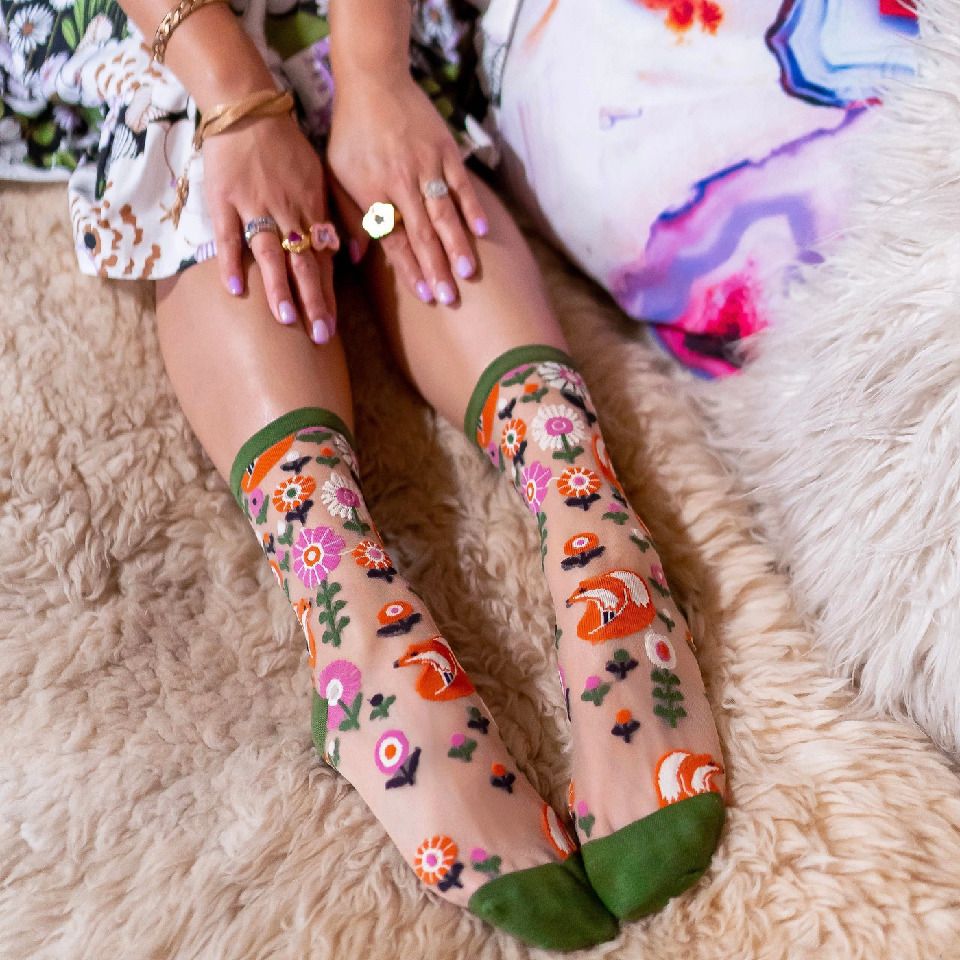 Legs on a fluffy rug wearing sheer fox socks, hands resting on knees with rings and a floral dress.