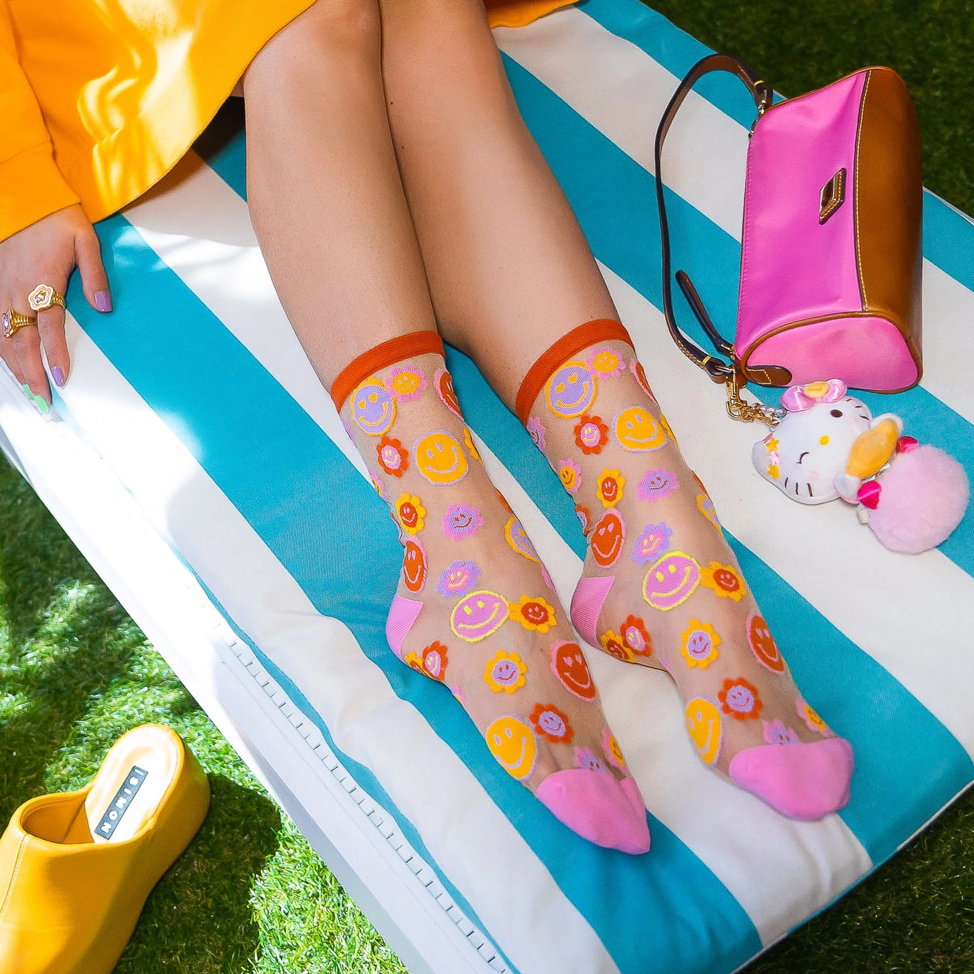 Legs crossed on a blue and white striped lounge chair wearing sheer smiley flower socks, next to a pink bag and cat plush.