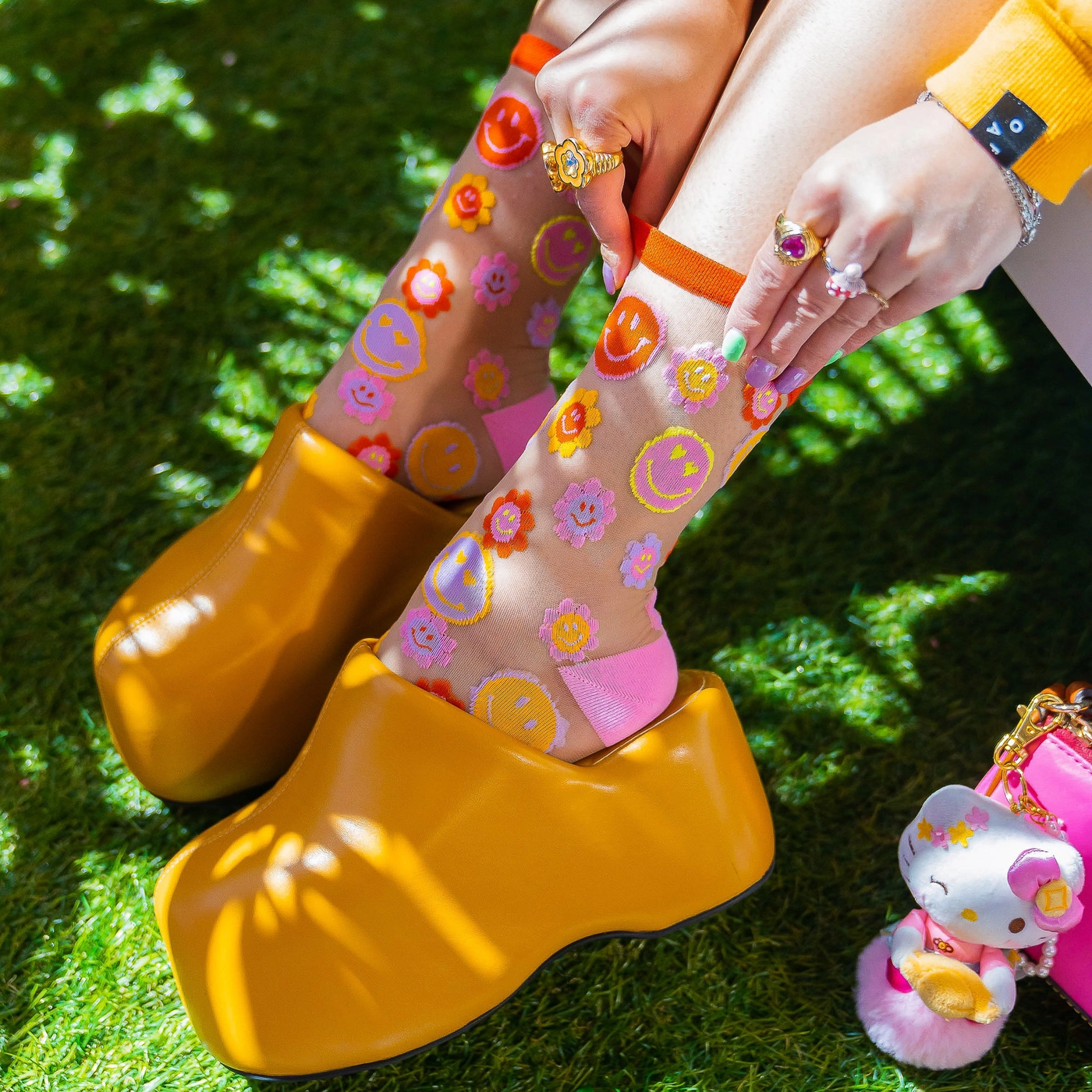 Close up of legs wearing sheer smiley daisy socks and chunky yellow clog shoes, standing on bright green artificial grass.
