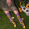 Person on grass wearing sheer sunflower socks with solid yellow toes, hand with painted nails resting on a lace dress.