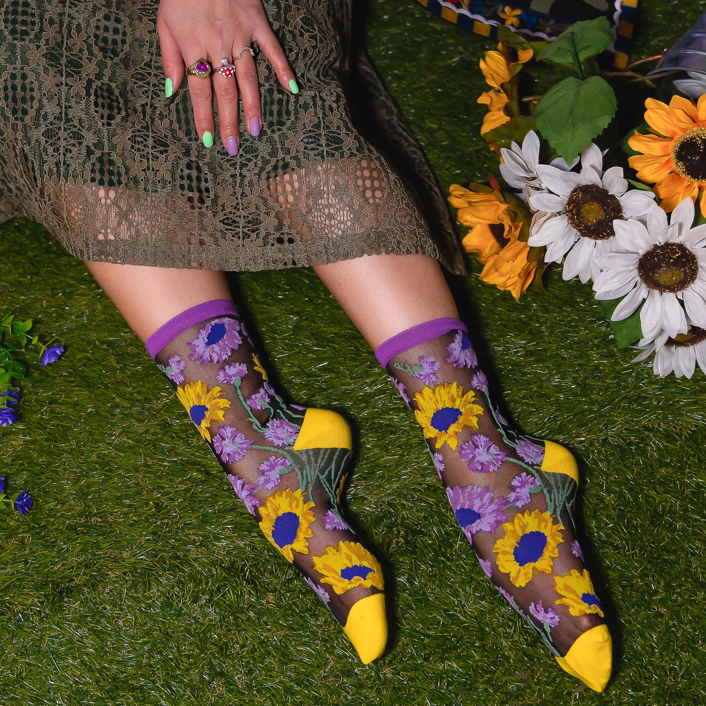 Person on grass wearing sheer sunflower socks with solid yellow toes, hand with painted nails resting on a lace dress.