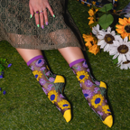 Person on grass wearing sheer sunflower socks with solid yellow toes, hand with painted nails resting on a lace dress.
