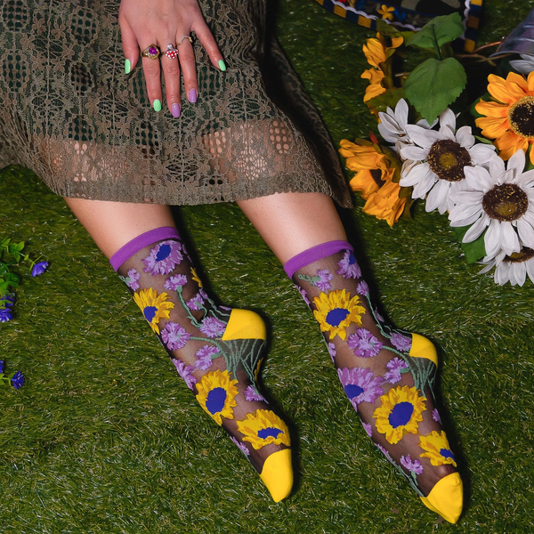 [lf] Person on grass wearing sheer sunflower socks with solid yellow toes, hand with painted nails resting on a lace dress.