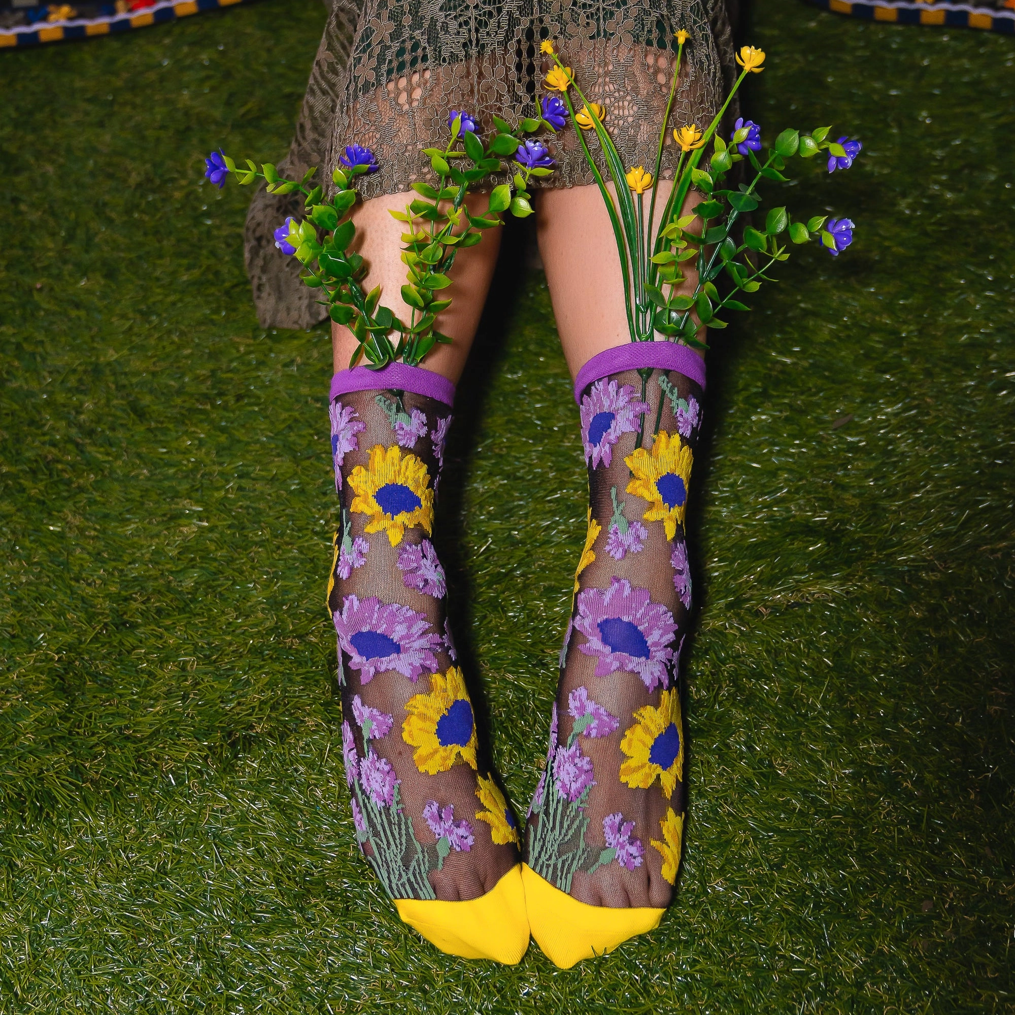 Legs on artificial grass wearing sheer floral socks, with fake purple and yellow flower stems tucked into the purple cuffs.