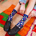 Person wearing sheer blue floral socks and black heeled loafers, sitting on a bright multicolored rug with hands resting.