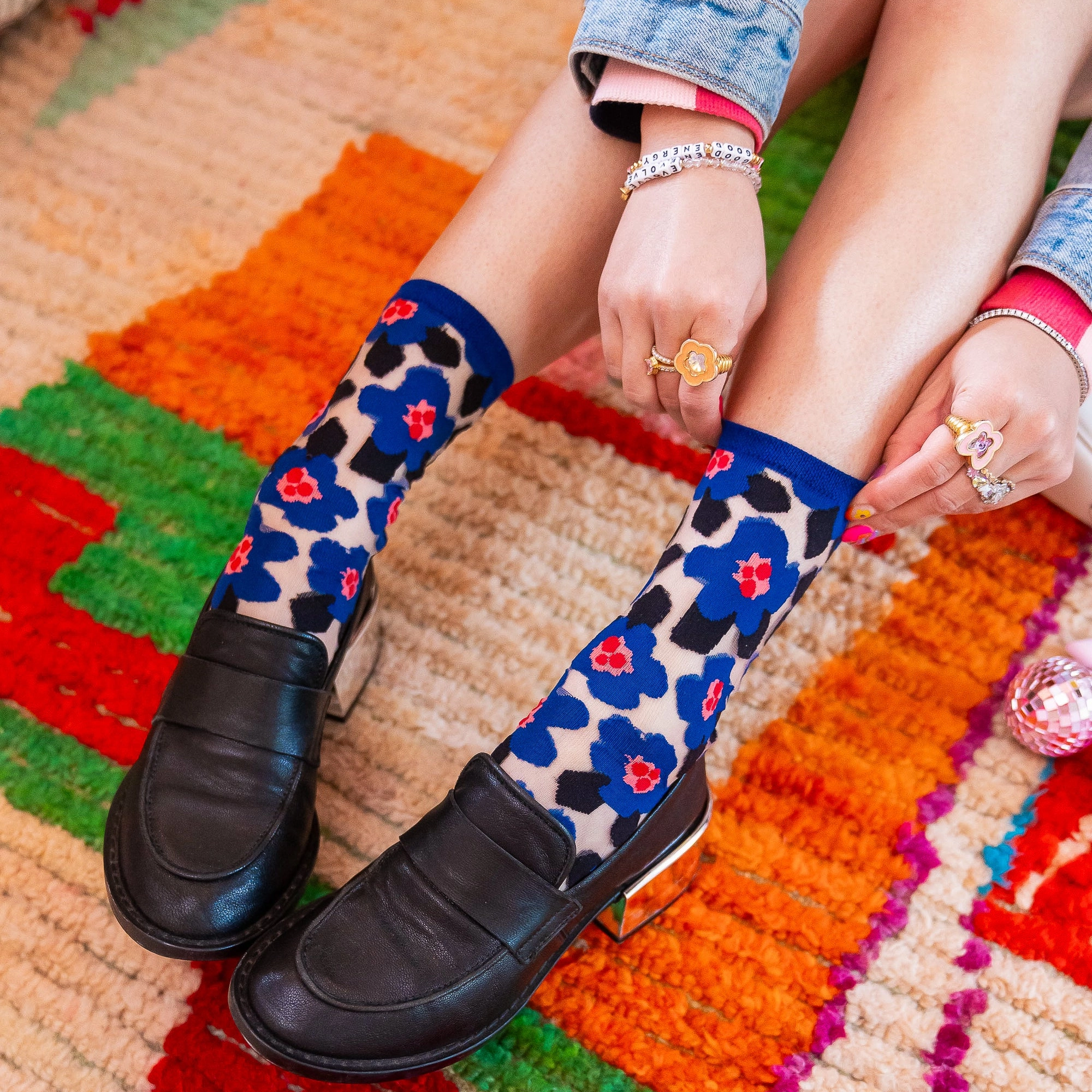 Person wearing sheer blue floral socks and black heeled loafers, sitting on a bright multicolored rug with hands resting.