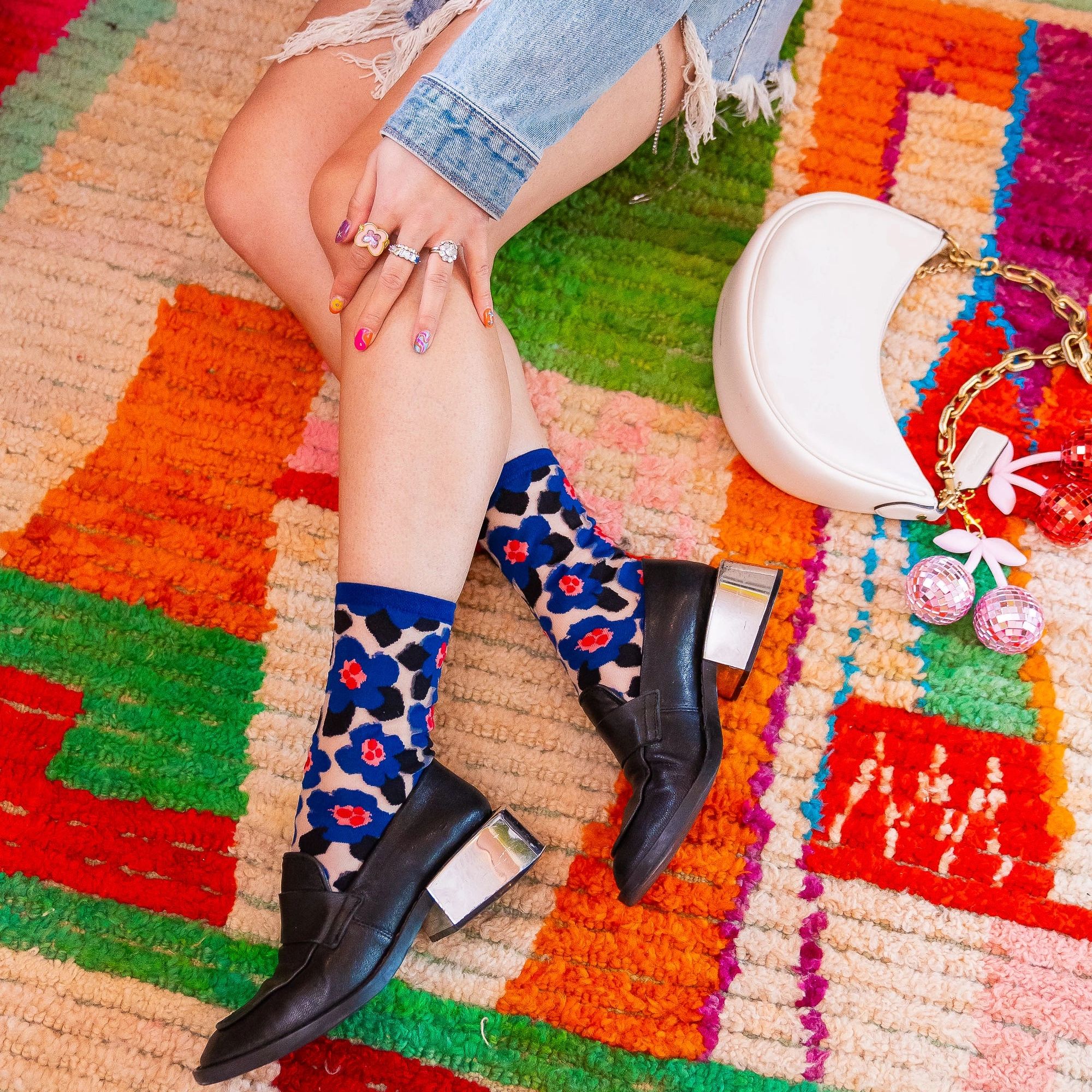 Crossed legs in sheer floral socks and black loafers on a colorful rug next to a white handbag and disco ball cherries.