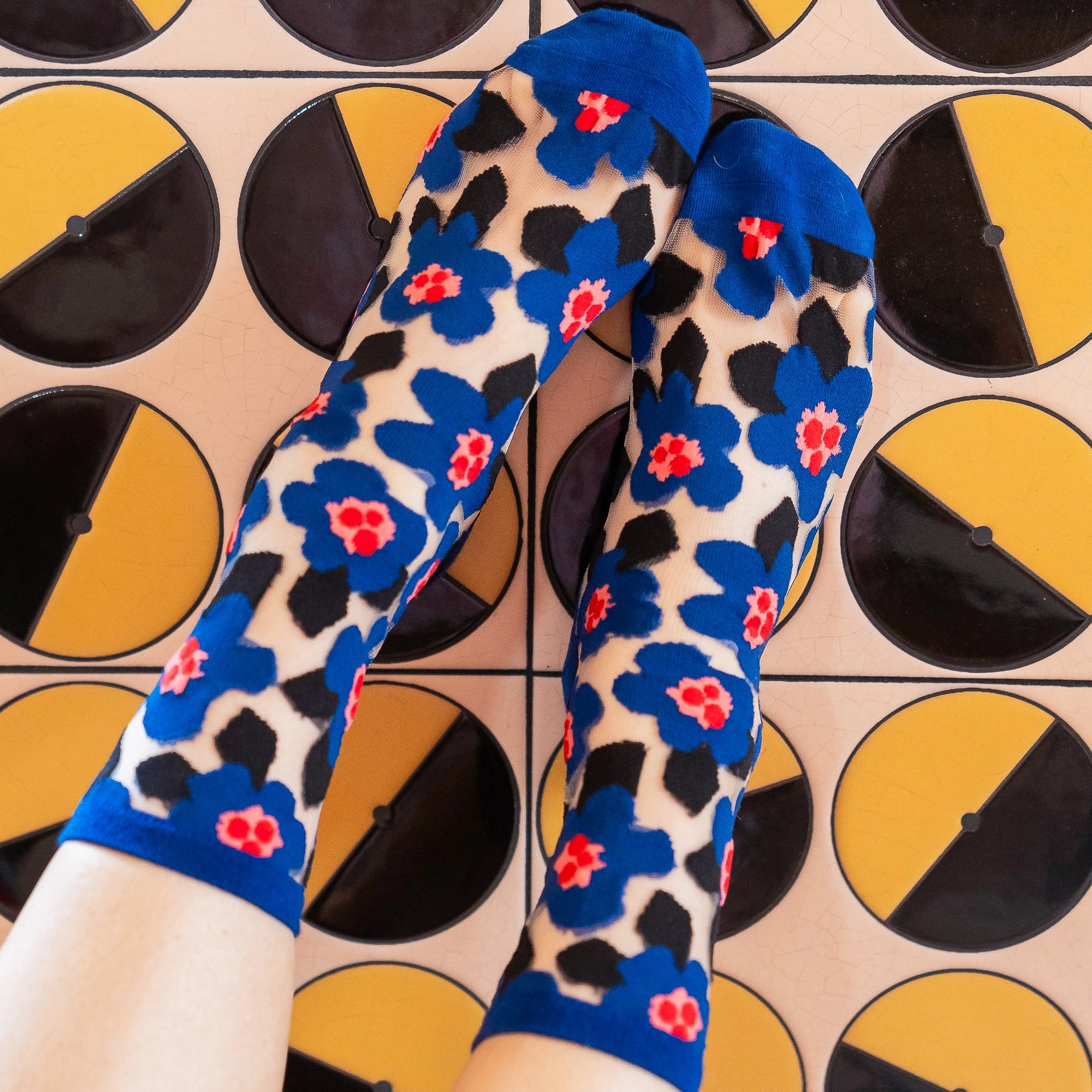 Point of view looking down at feet wearing sheer blue floral socks, standing against geometric black and yellow tiles.