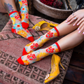 Person with jewelry pulling up a sheer floral sock on a patterned rug next to someone with a floral foot tattoo.