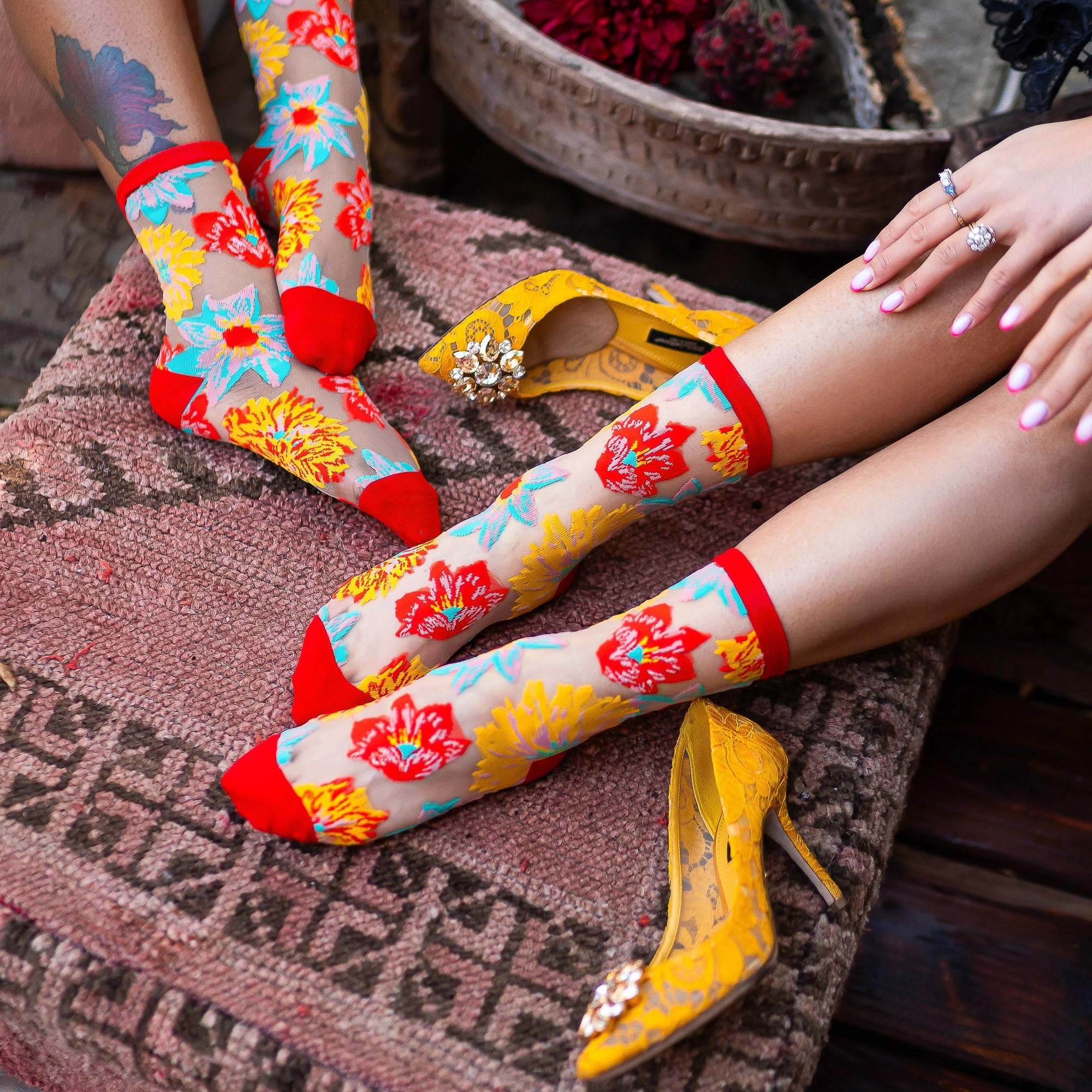 Person with jewelry pulling up a sheer floral sock on a patterned rug next to someone with a floral foot tattoo.