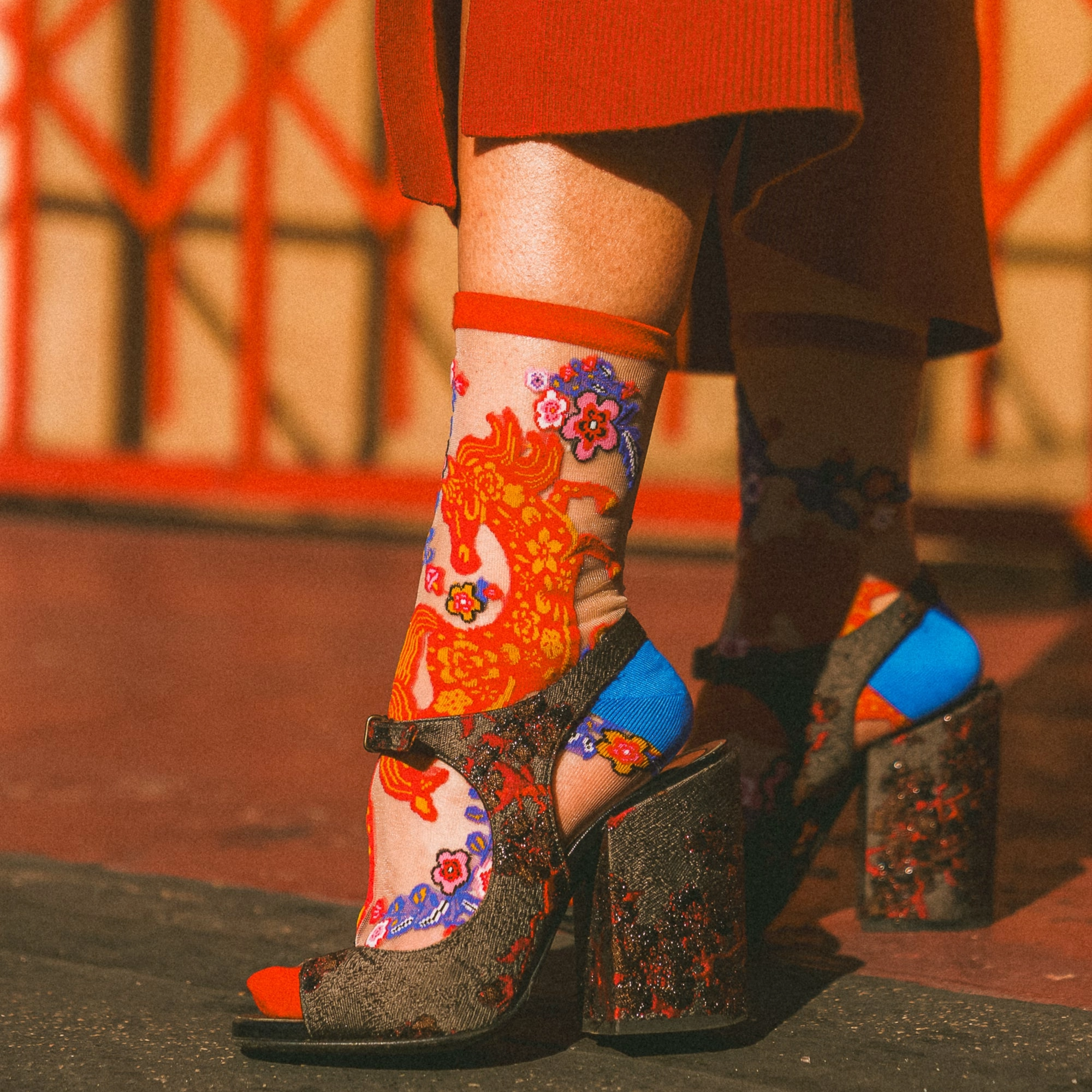 Close-up of hands adjusting the red cuff of a sheer floral horse sock, worn with dark sparkly open-toe block heels.