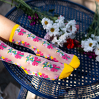 Legs crossed on a blue metal lattice table wearing sheer floral socks, surrounded by real white and purple daisies.