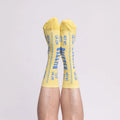 A pair of yellow crew socks with blue text reading 'BUTTER'  worn on a person's legs against a plain light gray background.
