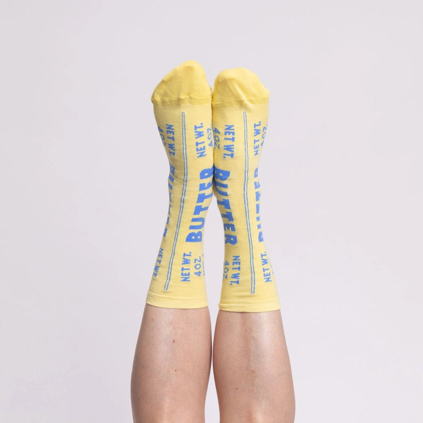 A pair of yellow crew socks with blue text reading 'BUTTER'  worn on a person's legs against a plain light gray background.