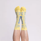 A pair of yellow crew socks with blue text reading 'BUTTER'  worn on a person's legs against a plain light gray background.