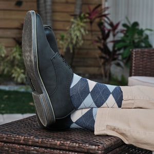 Blue Argyle dress socks with cream and grey diamond pattern shown above black leather boots, with khaki pants resting on a wicker ottoman in a tropical backyard. [lf]